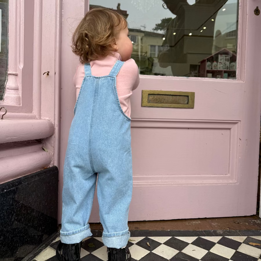 Child in blue overalls standing in front of a pink door with a checkered floor.